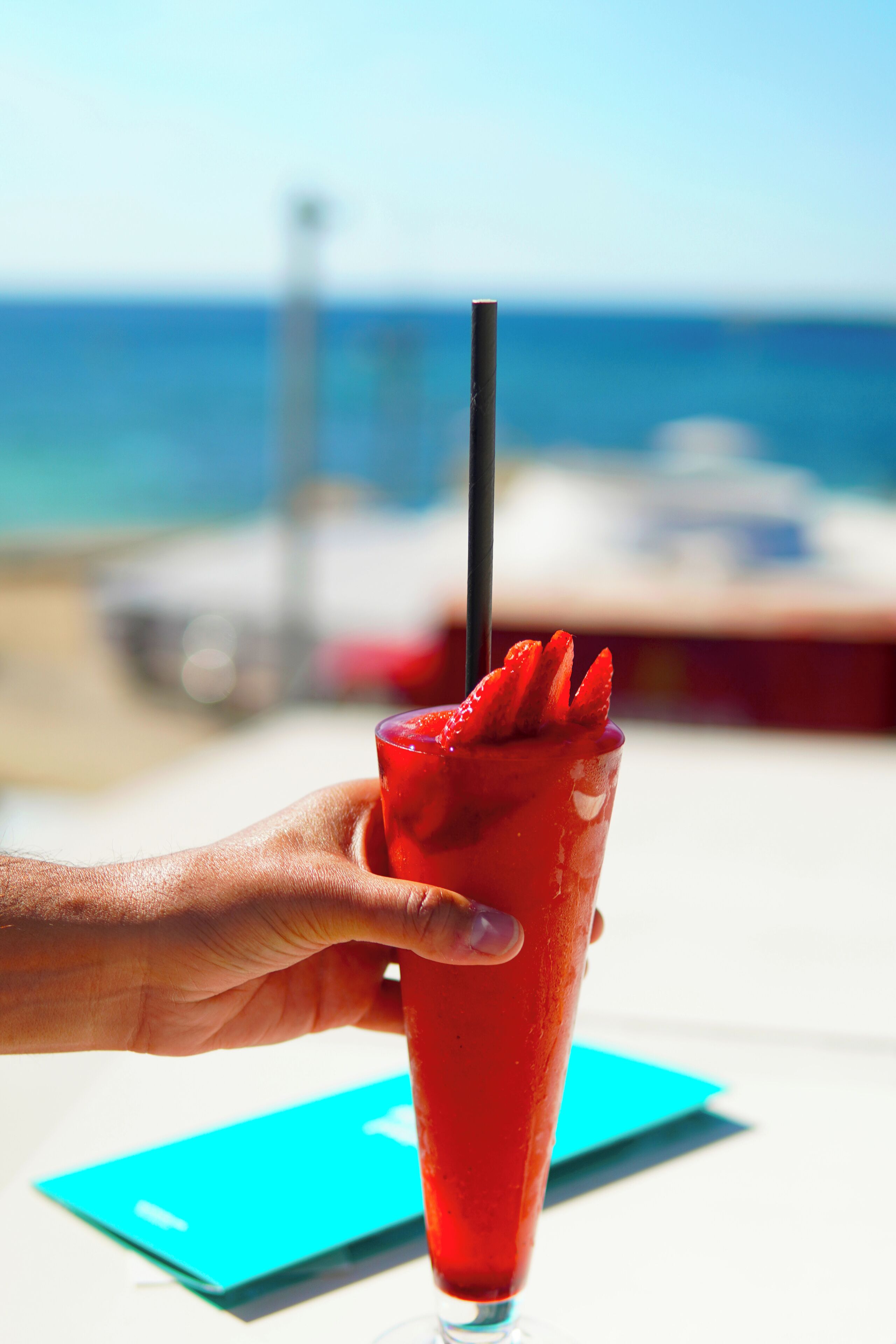 Une main tenant un verre de boisson rouge avec une paille, sur fond d’une plage et d’un océan flous.