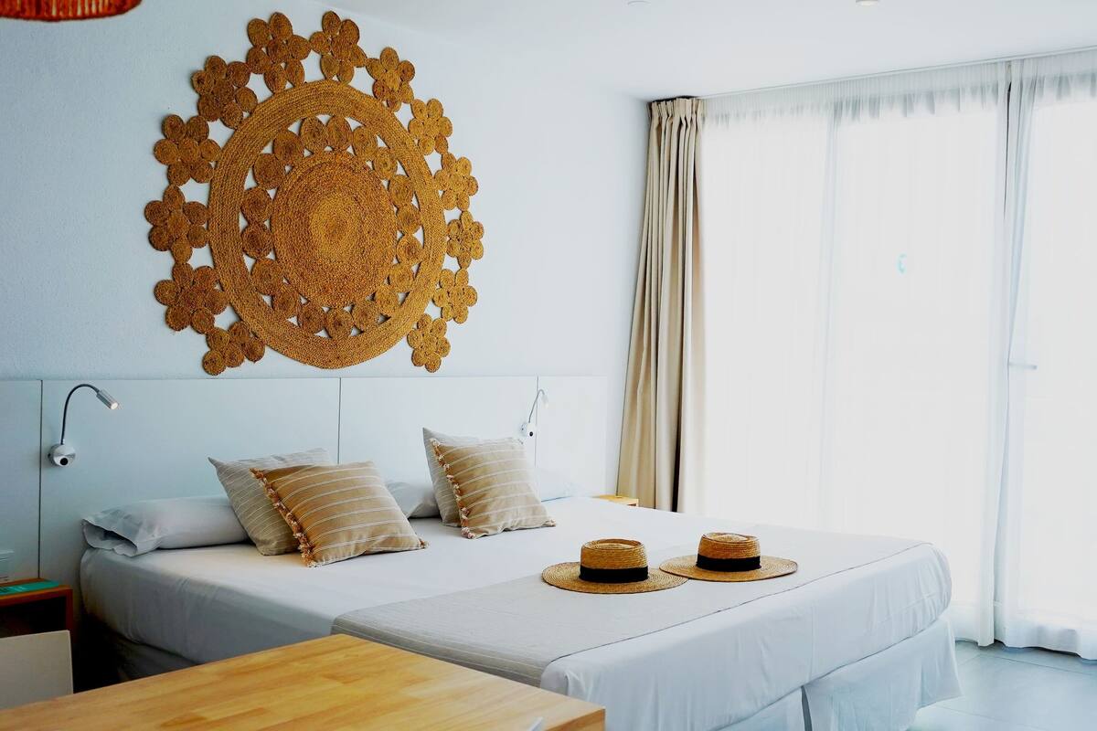 Een hotelkamer met twee bedden, een groot decoratief wandobject, een nachtkastje en uitzicht op de omgeving door grote ramen.