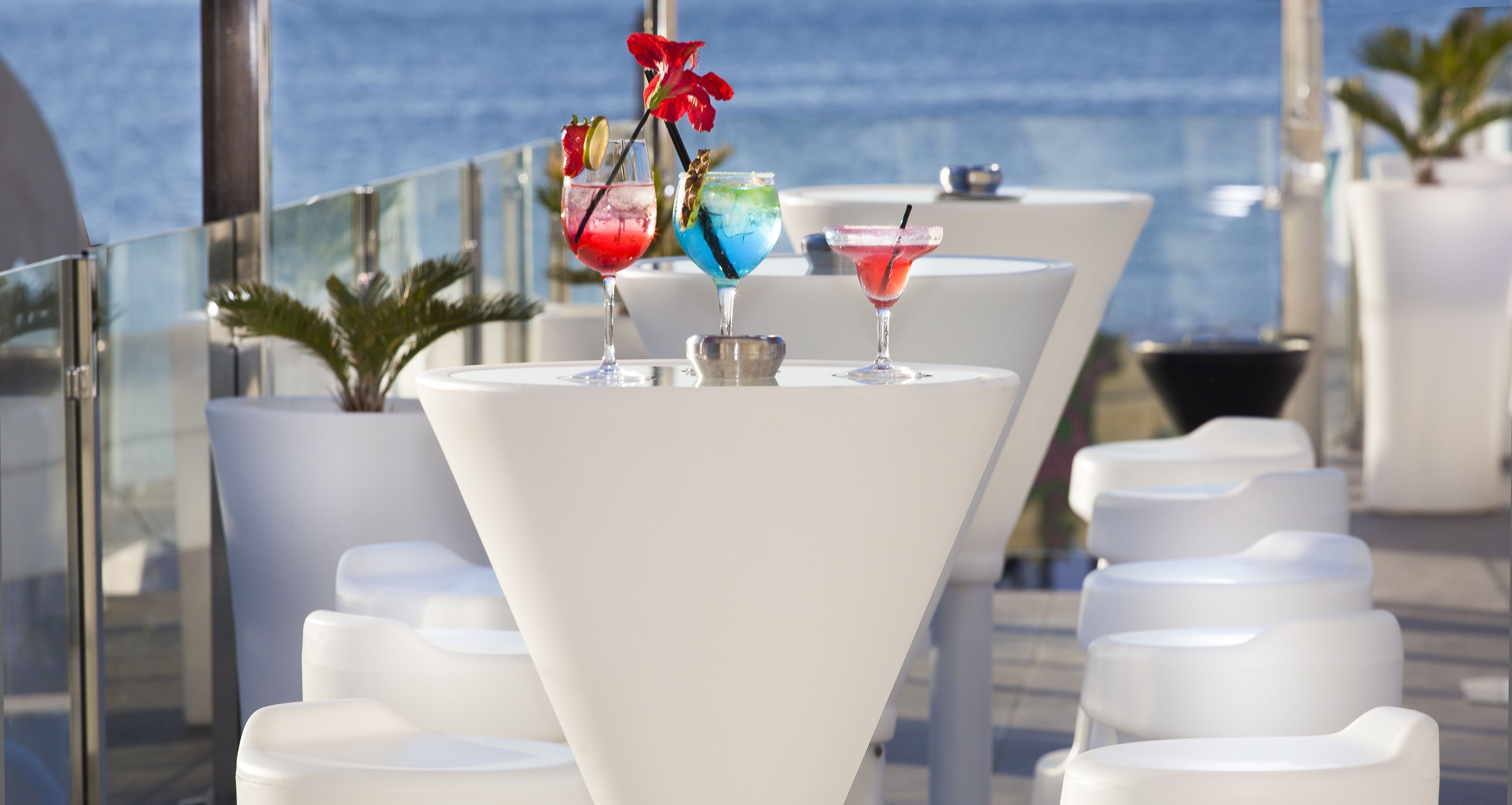 A rooftop bar with white tables and chairs, three cocktails with garnishes, and a view of the ocean.