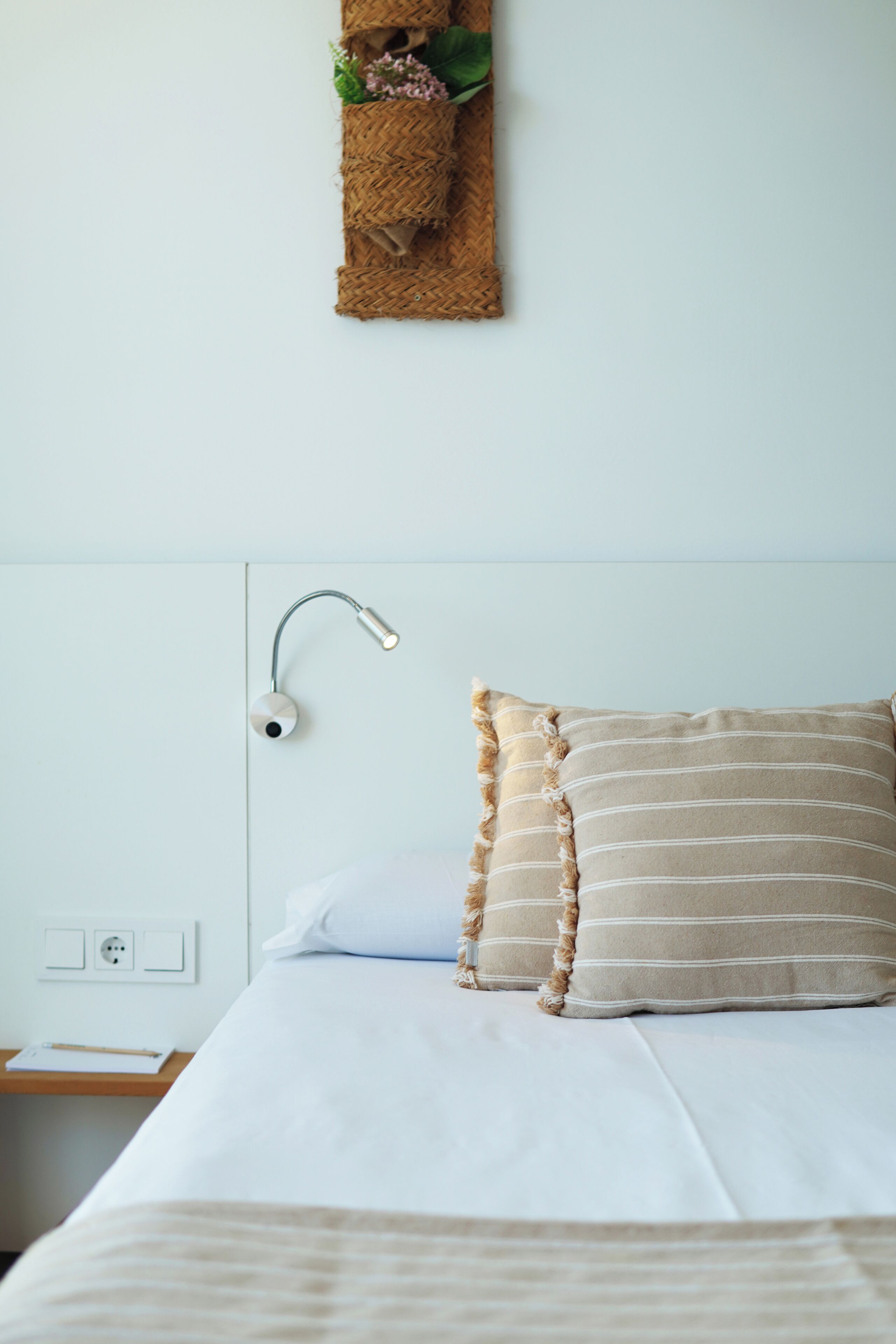 Ein ordentlich bezogenes Bett mit weißer Bettwäsche und einem gestreiften Kissen, ein Nachttisch mit Lampe und eine Wandregal mit Dekoration.