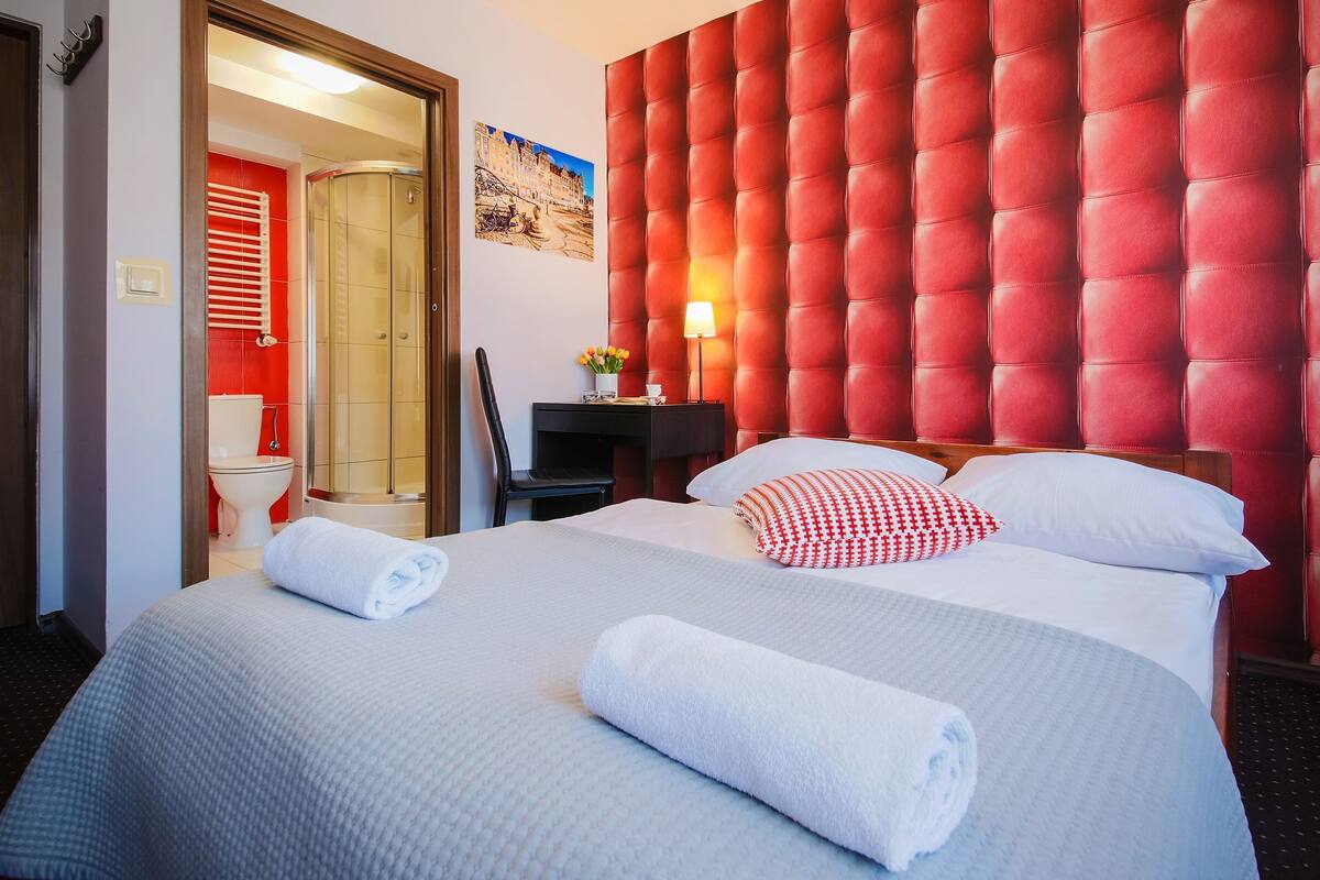 A hotel room with two beds, a red upholstered headboard, a painting on the wall, and a bathroom visible through an open door.