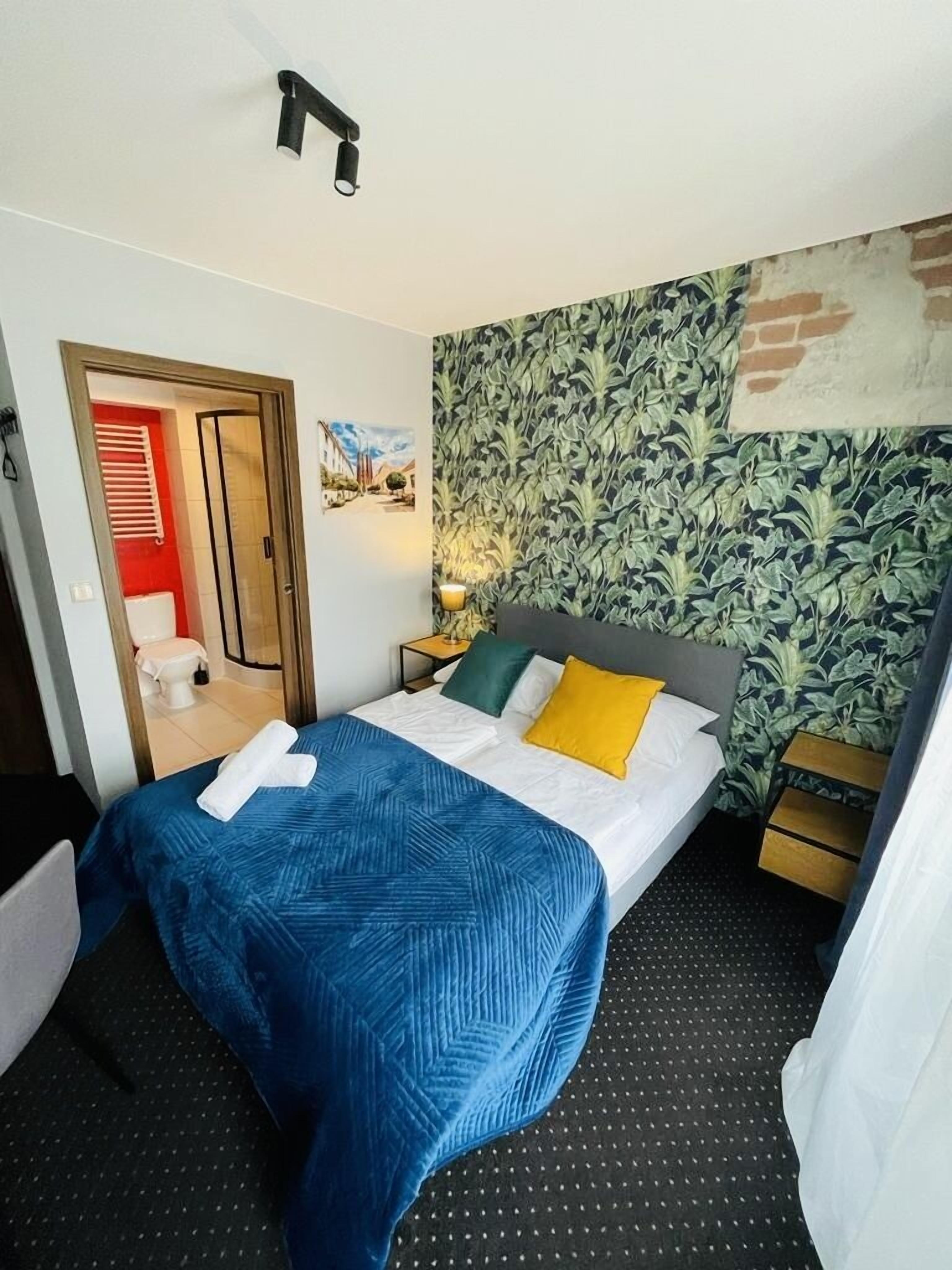A hotel room with a bed, a nightstand, a wall with a leafy pattern, and a door leading to a bathroom.