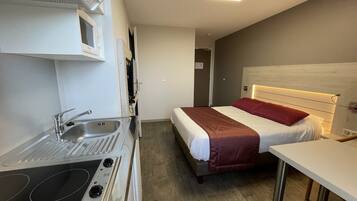 Una habitación de hotel compacta con kitchenette, una cama con manta roja y una mesita de noche.