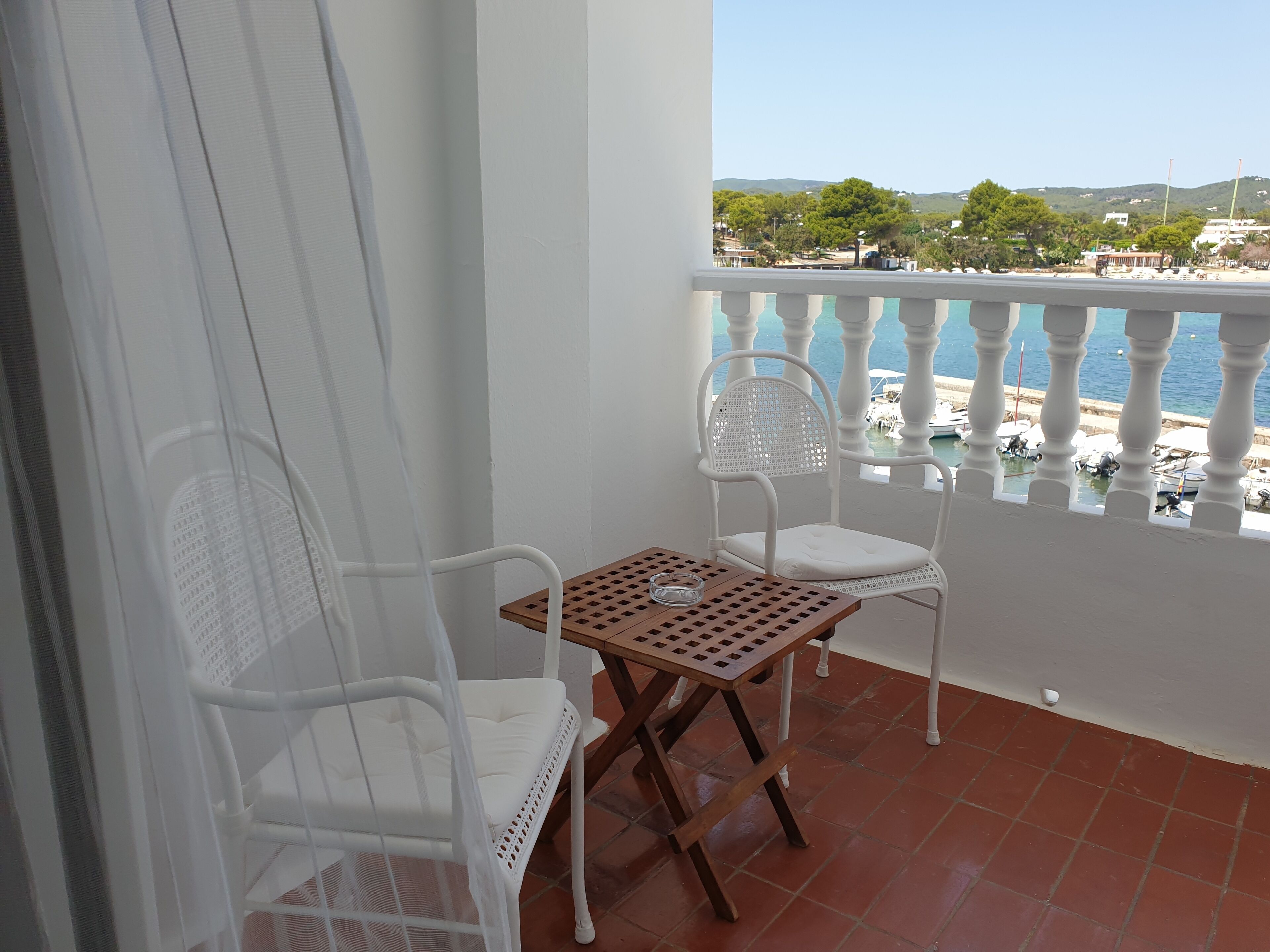 A balcony with two white wicker chairs, a wooden table, and a view of the sea and buildings.