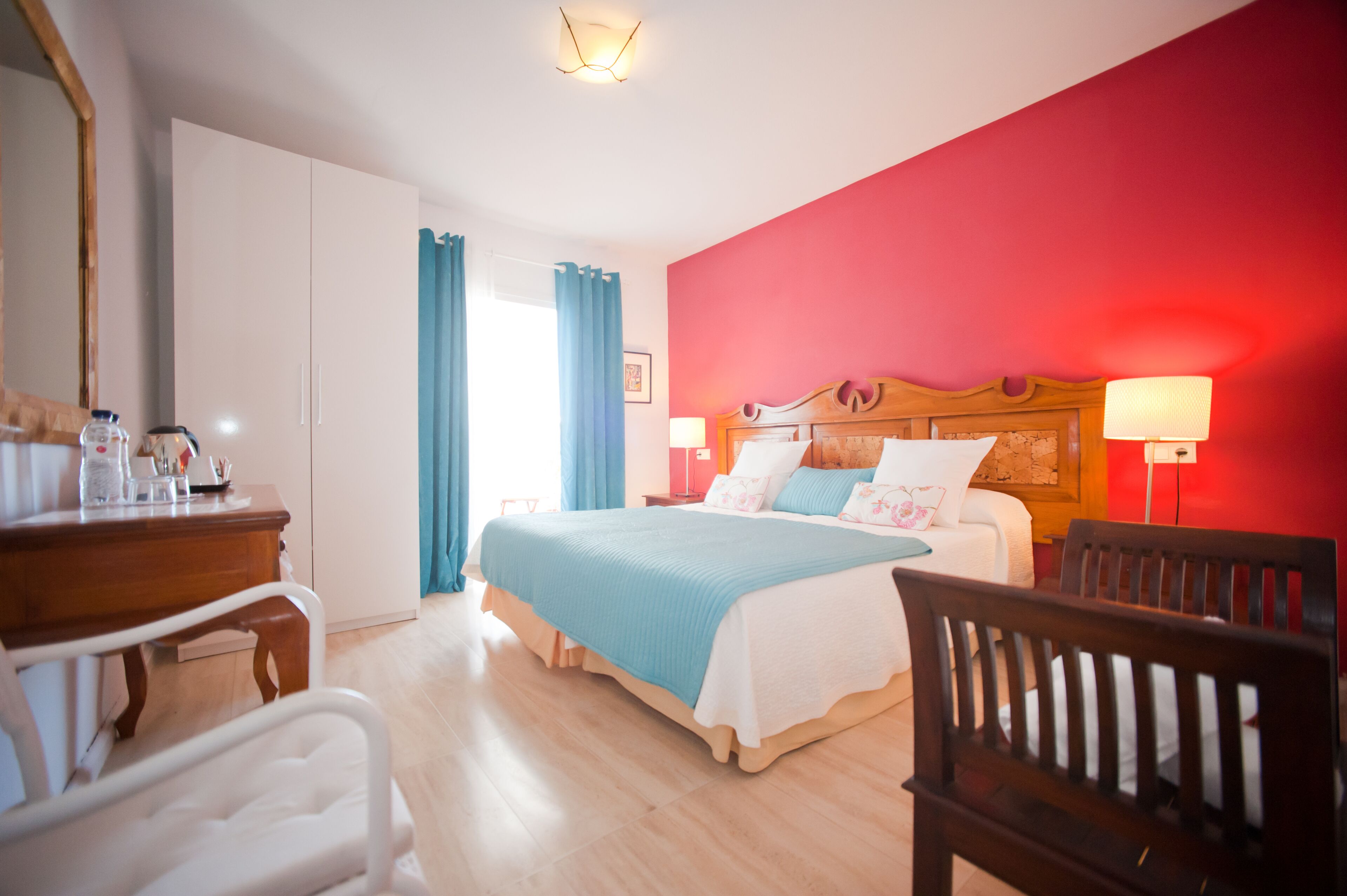A hotel room with a bed, bedside lamps, a wooden chair, a desk with a mirror, and a wardrobe.