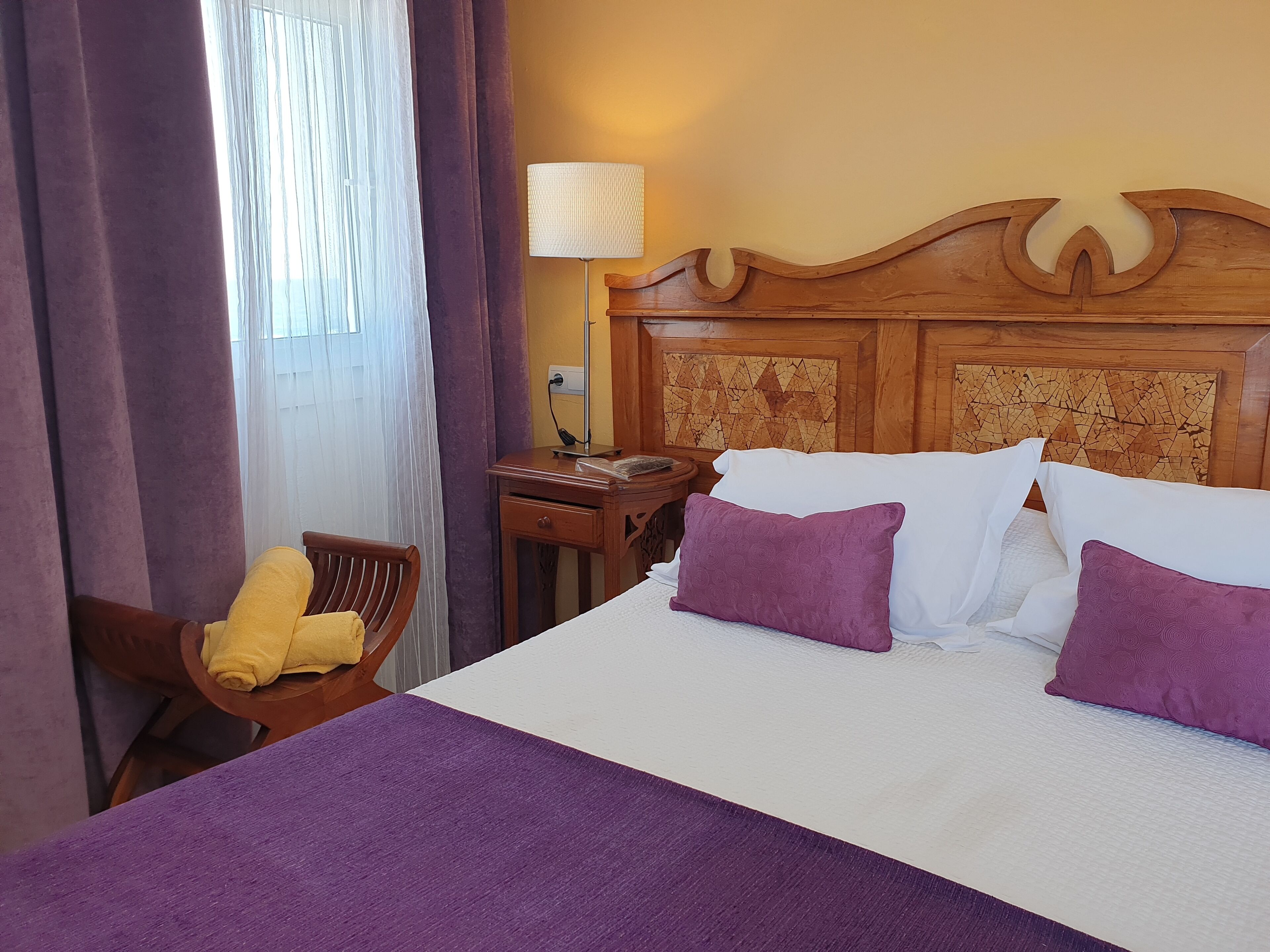 Una habitación de hotel con cabecera de madera, cama con ropa de cama blanca y morada, mesita de noche de madera con una lámpara y una silla con una toalla amarilla.