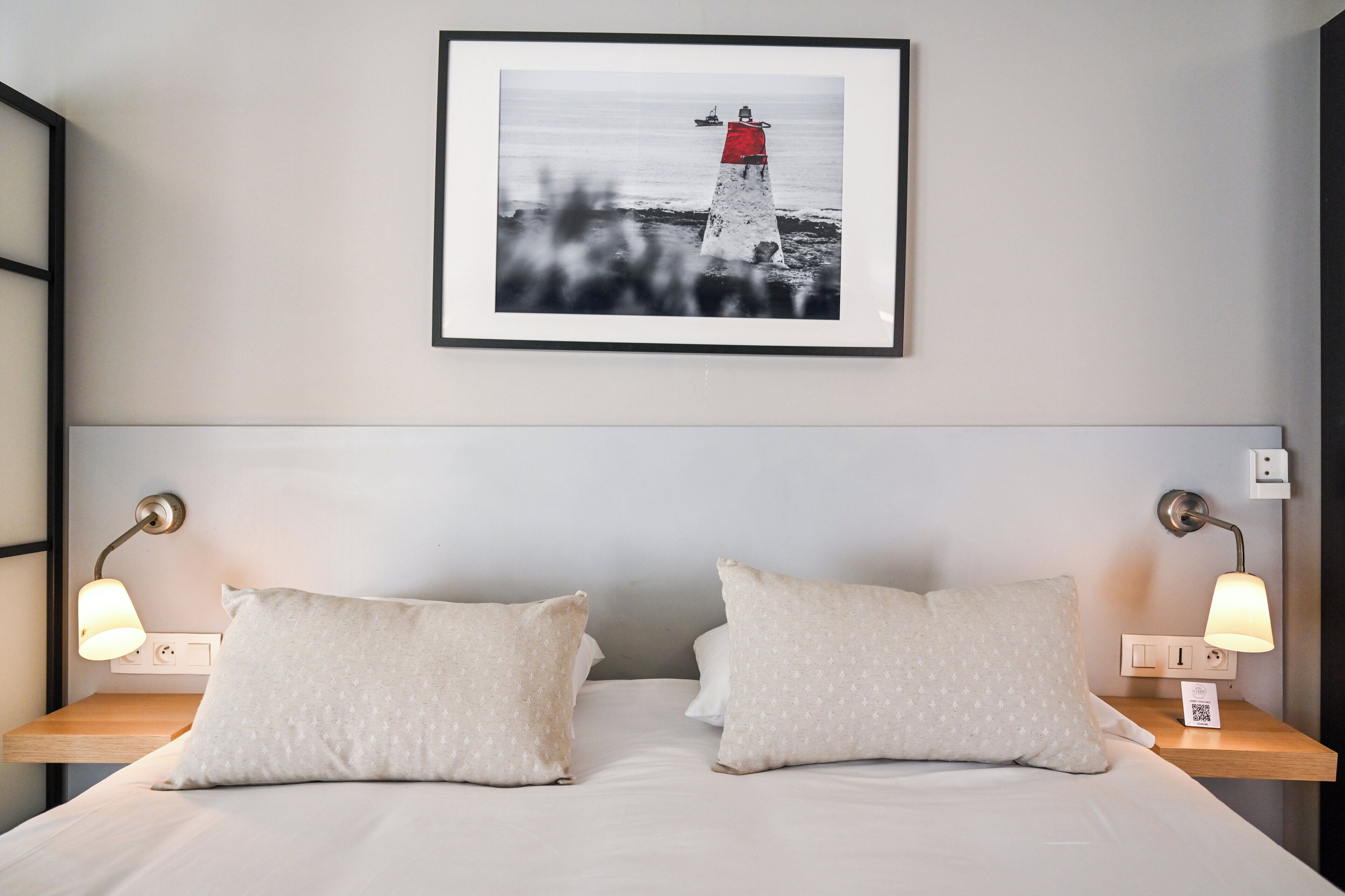 Una cama bien hecha con dos almohadas y dos lámparas de noche, un cuadro con un faro en la pared y una mesita de noche con un enchufe.