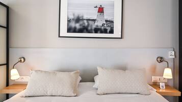 Una cama bien hecha con dos almohadas y dos lámparas de noche, un cuadro con un faro en la pared y una mesita de noche con un enchufe.