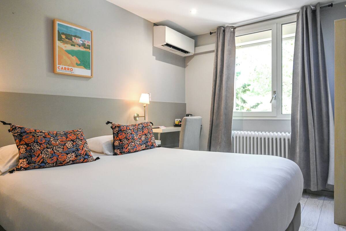 A hotel room with a bed, a desk, an air conditioner, and a window with curtains.
