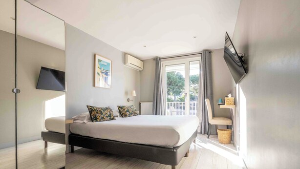 A modern bedroom with a large bed, a TV mounted on the wall, and a balcony with a view of trees. - Hôtel la Tuilière (Carry-le-Rouet)