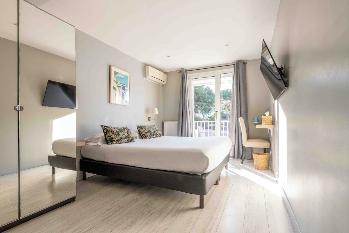 A modern bedroom with a large bed, a TV mounted on the wall, and a balcony with a view of trees.