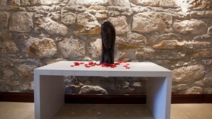 Une table blanche sur laquelle repose une sculpture, entourée de pétales rouges, le tout se détachant sur un mur de pierre.