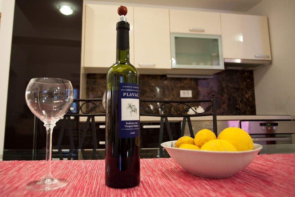 A bottle of Plavac wine, a glass, and a bowl of lemons on a table in a kitchen.