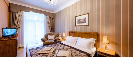 A hotel room with a large bed, a wooden headboard, a television on a wooden cabinet, a nightstand with a lamp, and a window with curtains.