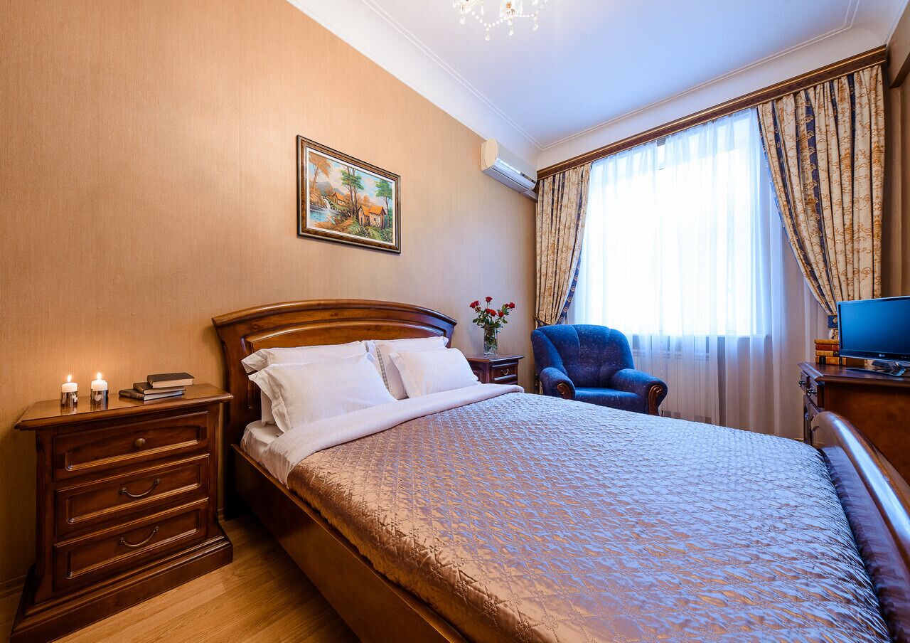 A neatly arranged bedroom with a large bed, a bedside table with a lamp, a chair, and a painting on the wall.