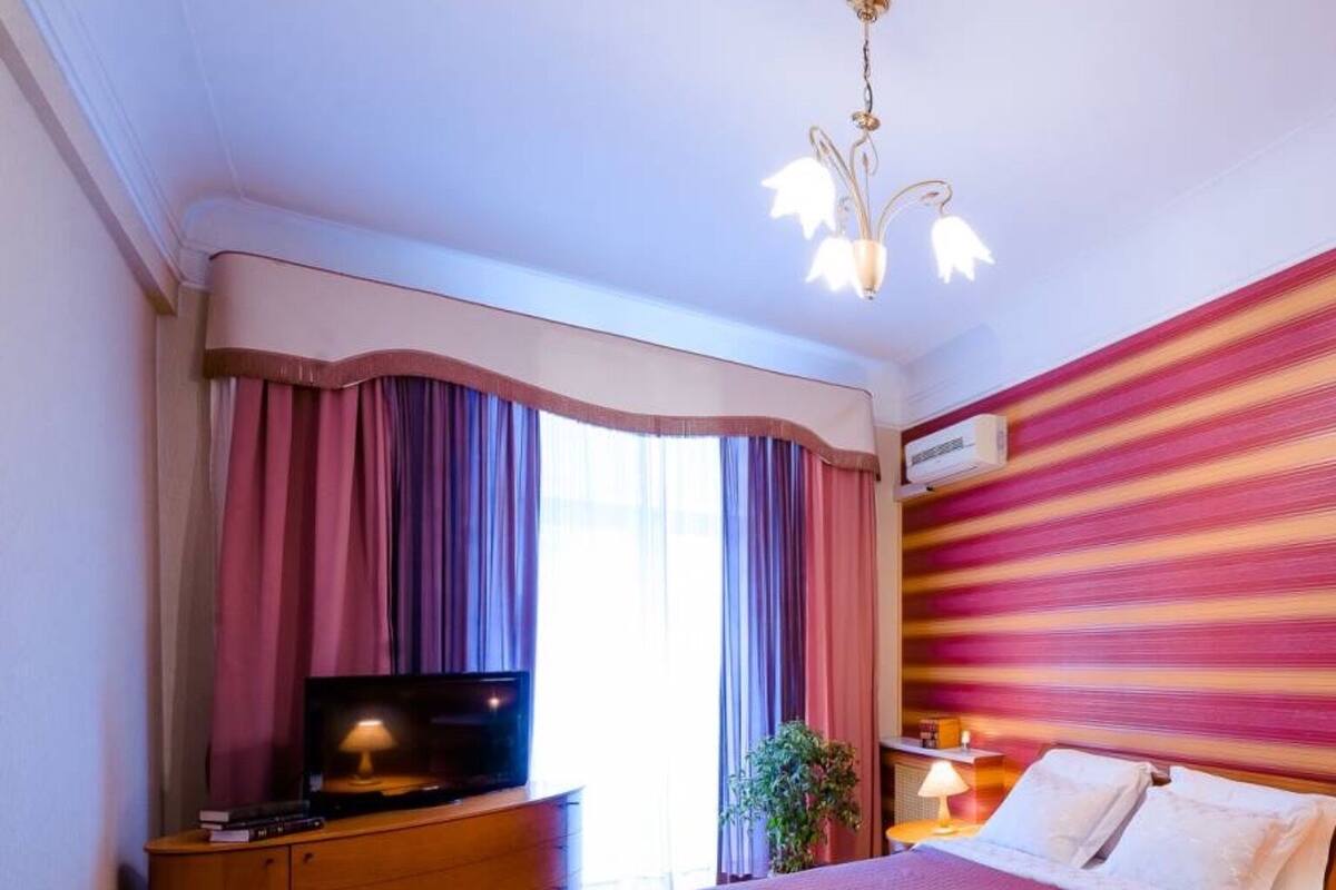Quarto com cama, televisão sobre um armário de madeira, mesa de cabeceira com abajur e janela com cortinas.
