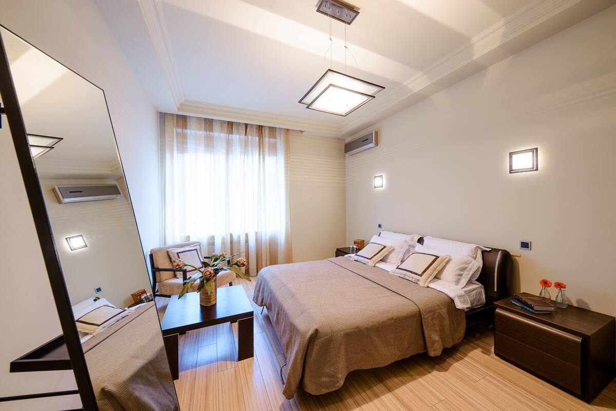 Quarto de hotel com cama, mesas de cabeceira, escrivaninha, cadeira, espelho e janela com cortinas.