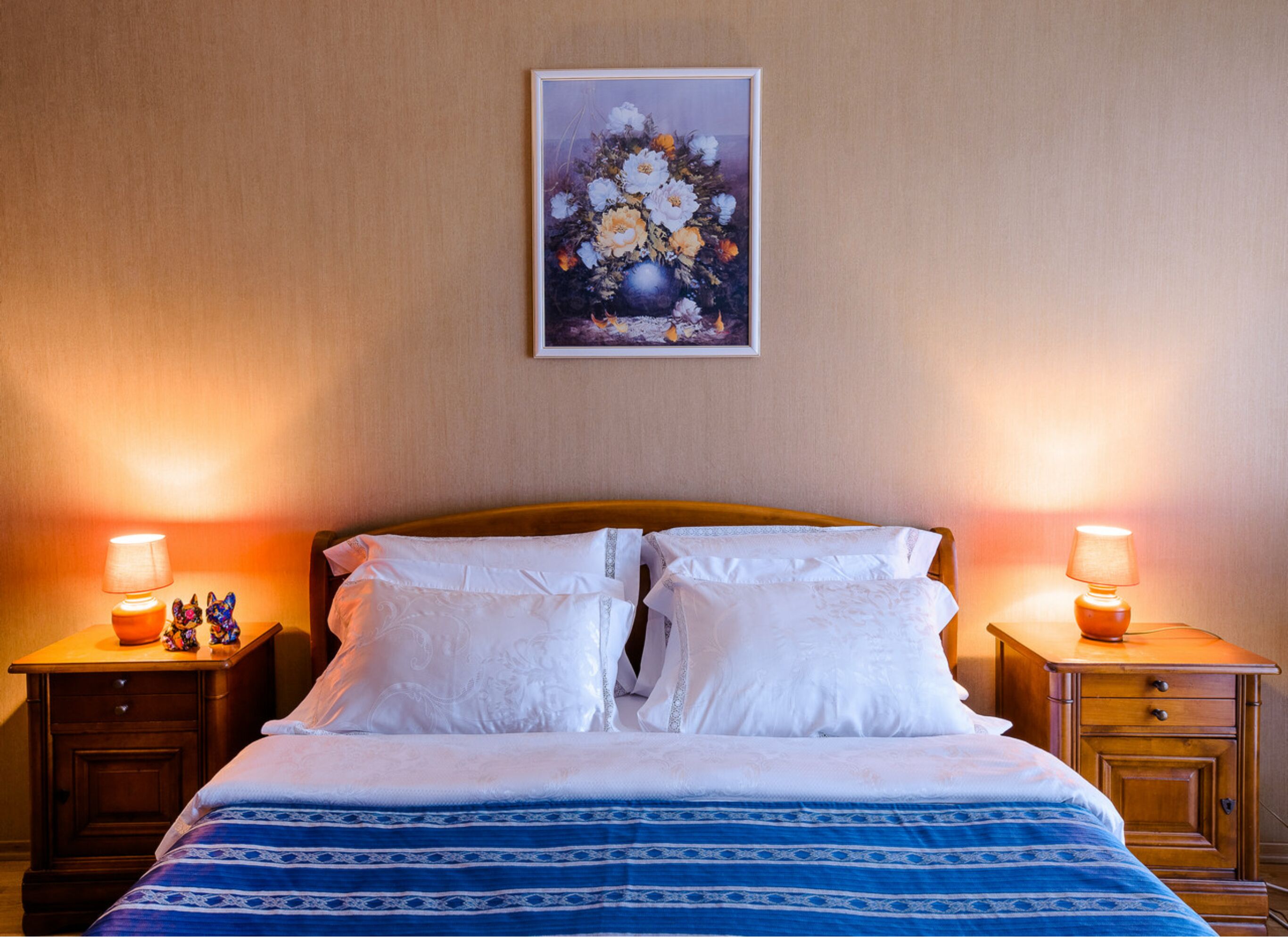 A neatly made bed with white linens and a blue patterned bedspread, flanked by two wooden nightstands with lamps and figurines, in a room with a beige wall and a framed floral artwork above the bed.