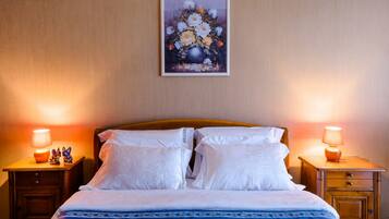 A neatly made bed with white linens and a blue patterned bedspread, flanked by two wooden nightstands with lamps and figurines, in a room with a beige wall and a framed floral artwork above the bed.
