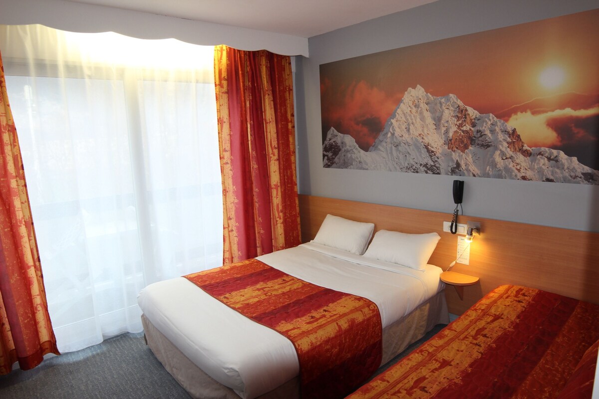 A hotel room with a bed, a bedside table, a lamp, and a painting of a mountain.