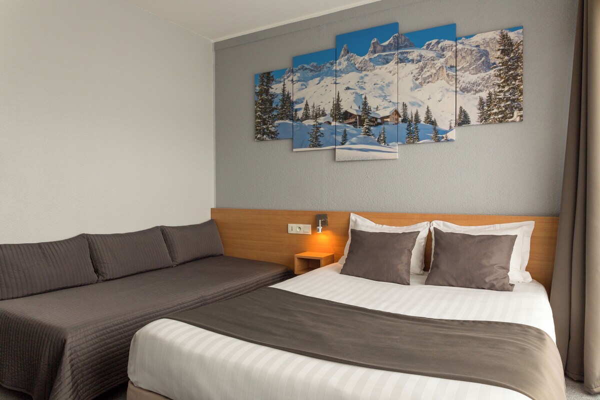 A hotel room with a bed, a sofa, a nightstand, and a wall-mounted painting of a snowy mountain landscape.