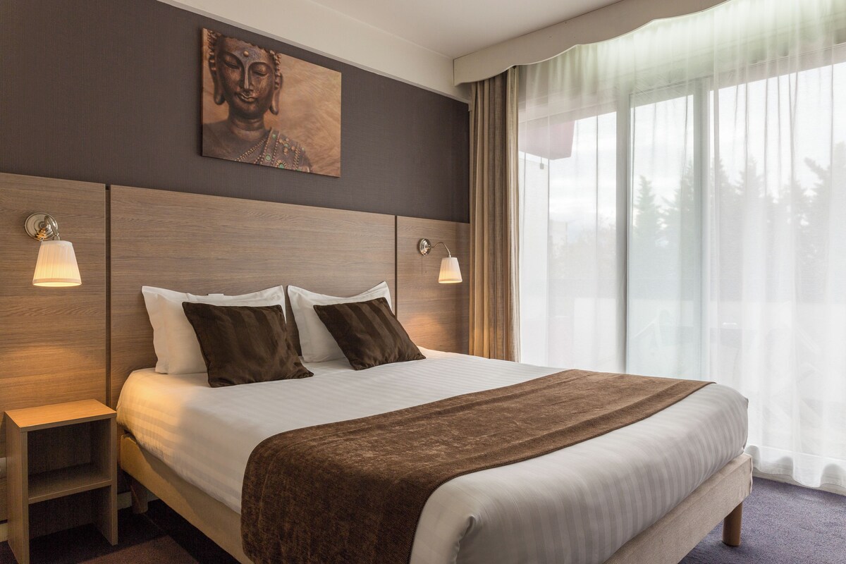 A hotel room with a large bed, two bedside tables with lamps, a wooden headboard, a Buddha painting, and a large window with curtains.
