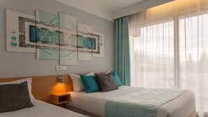Ett hotellrum med två sängar, en stor, abstrakt målning på väggen, ett nattduksbord med en lampa och utsikt över omgivningarna genom tunna gardiner.