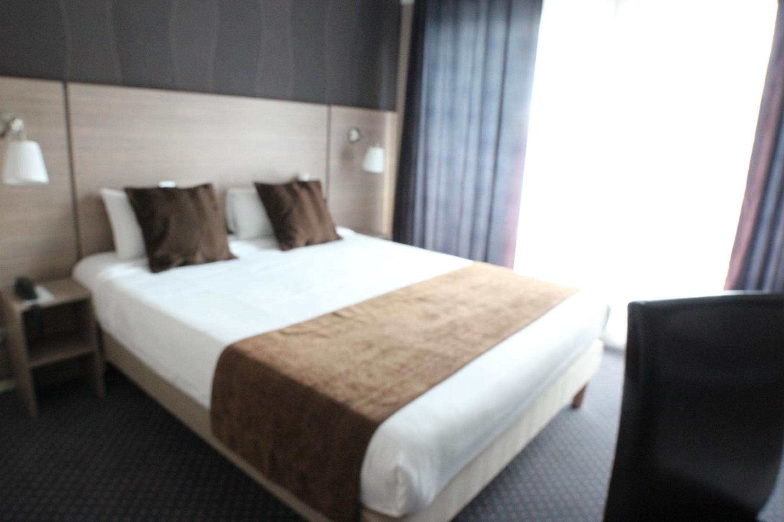 A hotel room with a large bed, a nightstand, a chair, and a headboard with wall-mounted lamps.