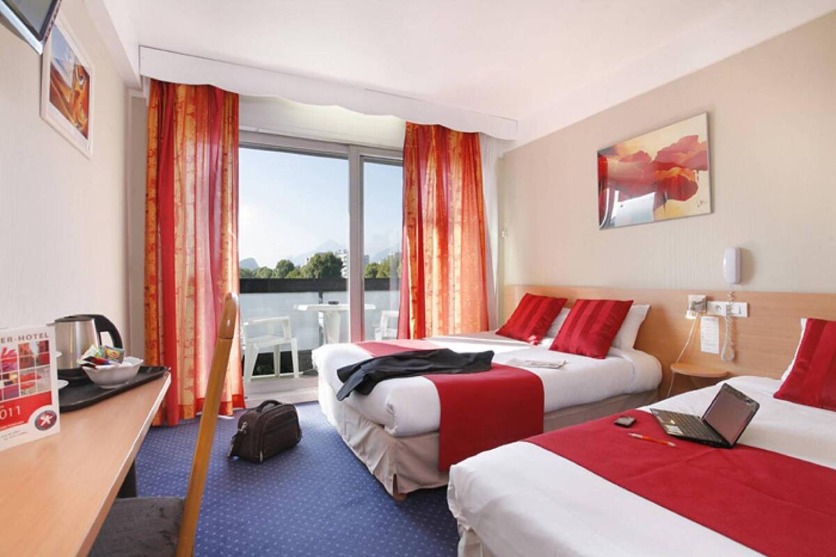 A hotel room with two beds, red bedding, a laptop on one bed, a desk with a coffee maker, and a view of the outdoors.