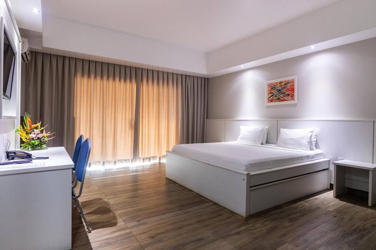 A hotel room with a bed, a desk, a chair, a small table, and a window with curtains.