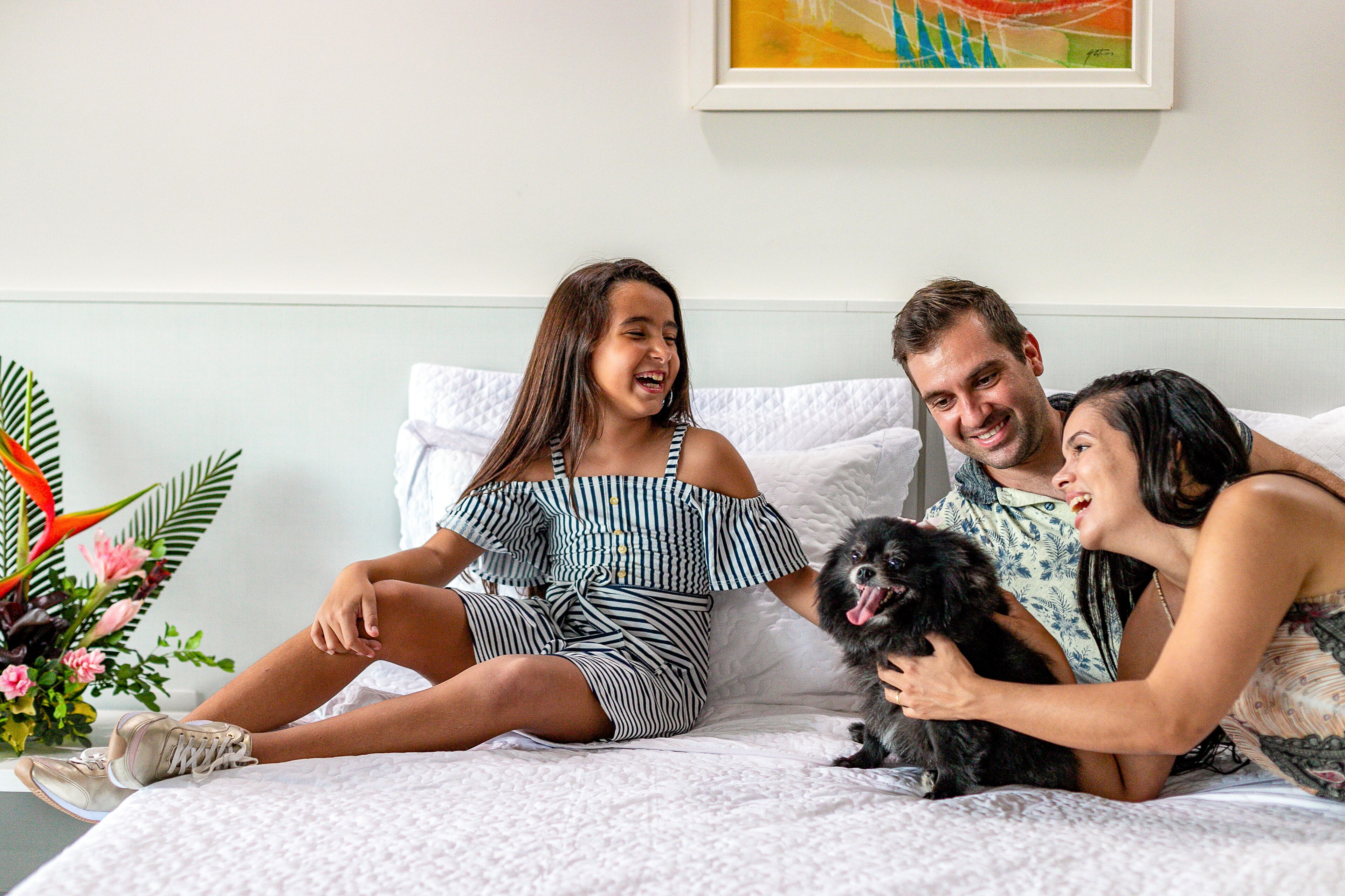 Uma família sentada em uma cama com um cão pequeno, cercada por uma parede branca e um quadro emoldurado.