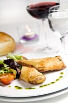 Un plato con un pastel enrollado, una rodaja de tomate y una ensalada, acompañado de una copa de vino tinto y un vaso de agua.