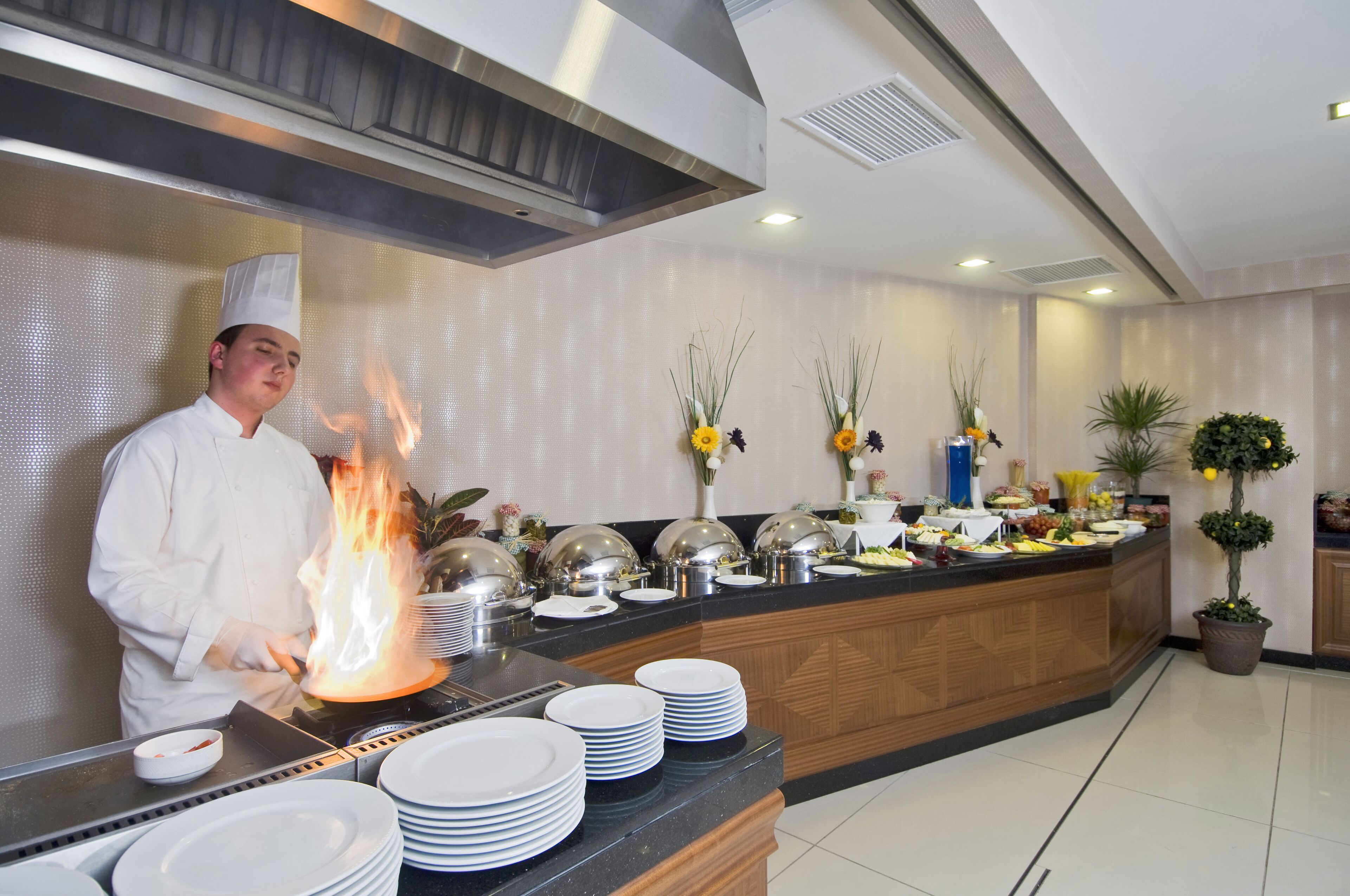 Un chef preparando un flambé en una cocina moderna con una barra de buffet, equipo de acero inoxidable y plantas decorativas.