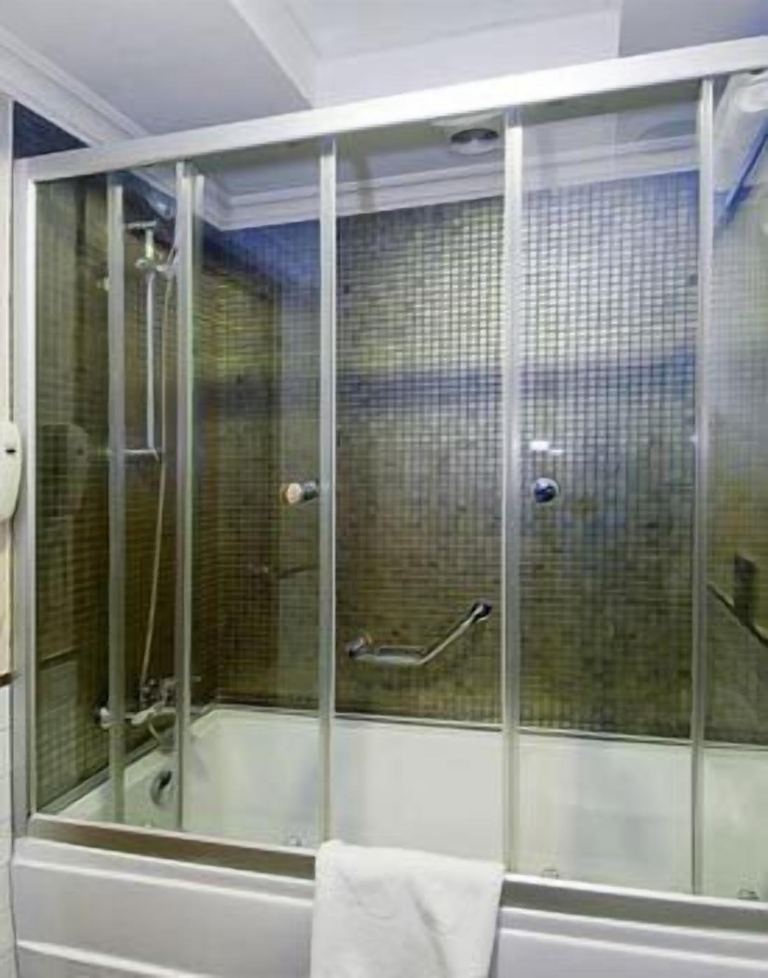 Ducha con paredes de vidrio, bañera blanca, cabezal de ducha manual y barra de apoyo.