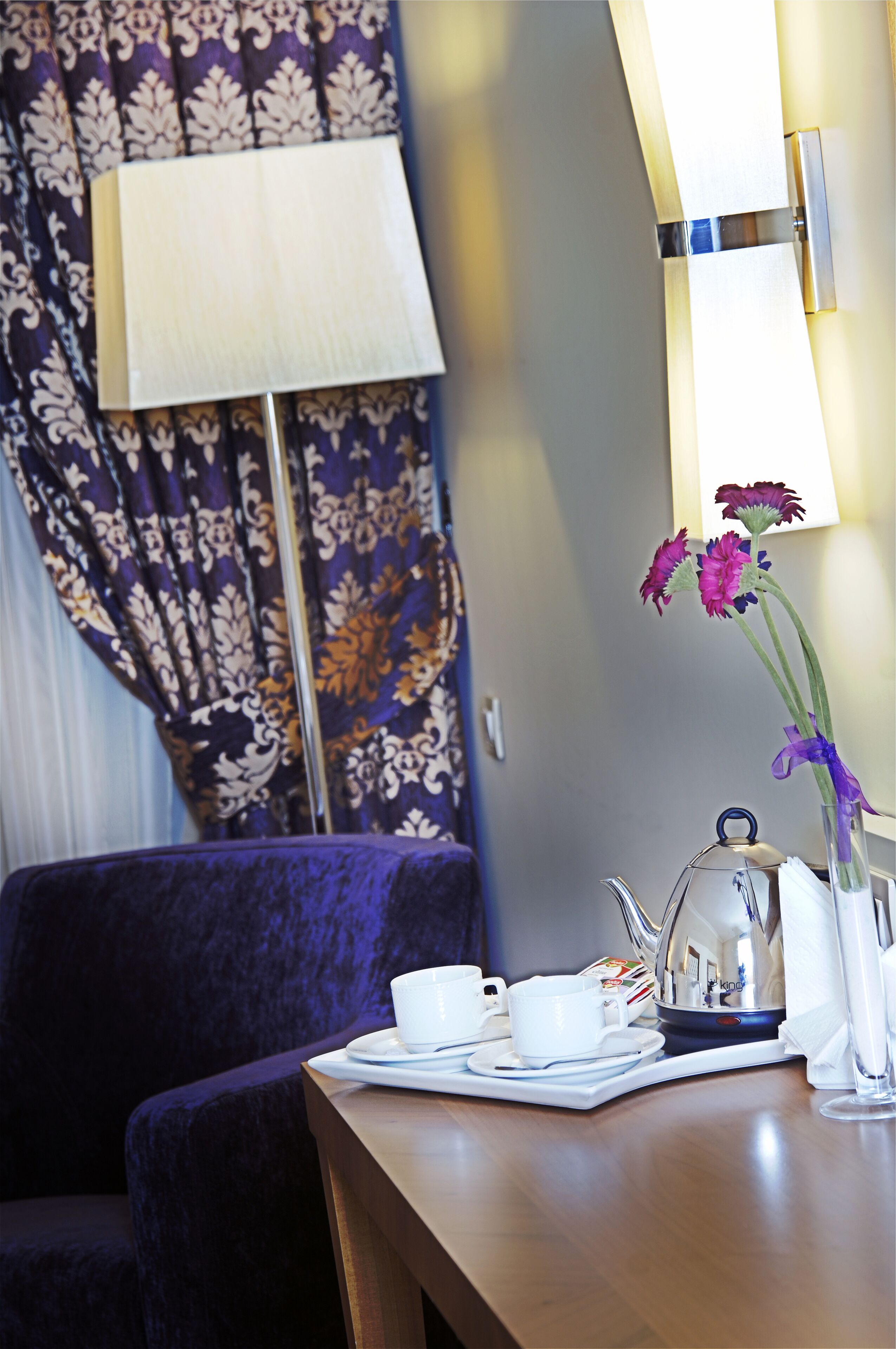 Un cuarto de hotel con una mesa preparada para tomar té, una lámpara y un florero con flores.