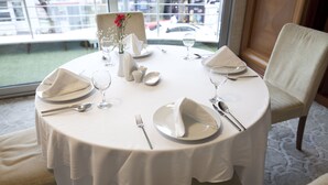 A round dining table set for four with white tablecloths, plates, cutlery, and glasses. A vase with flowers and a small salt and pepper set are on the table. The room has a large window with a view of the street outside. - Tugcu Hotel Select (Bursa)