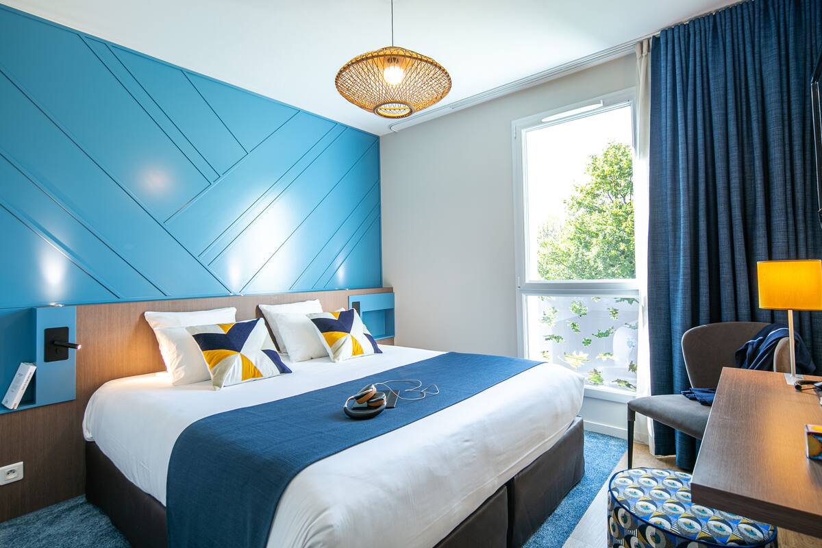 A modern hotel room with a large bed, a blue accent wall, a window with a view, and a small table with a lamp.