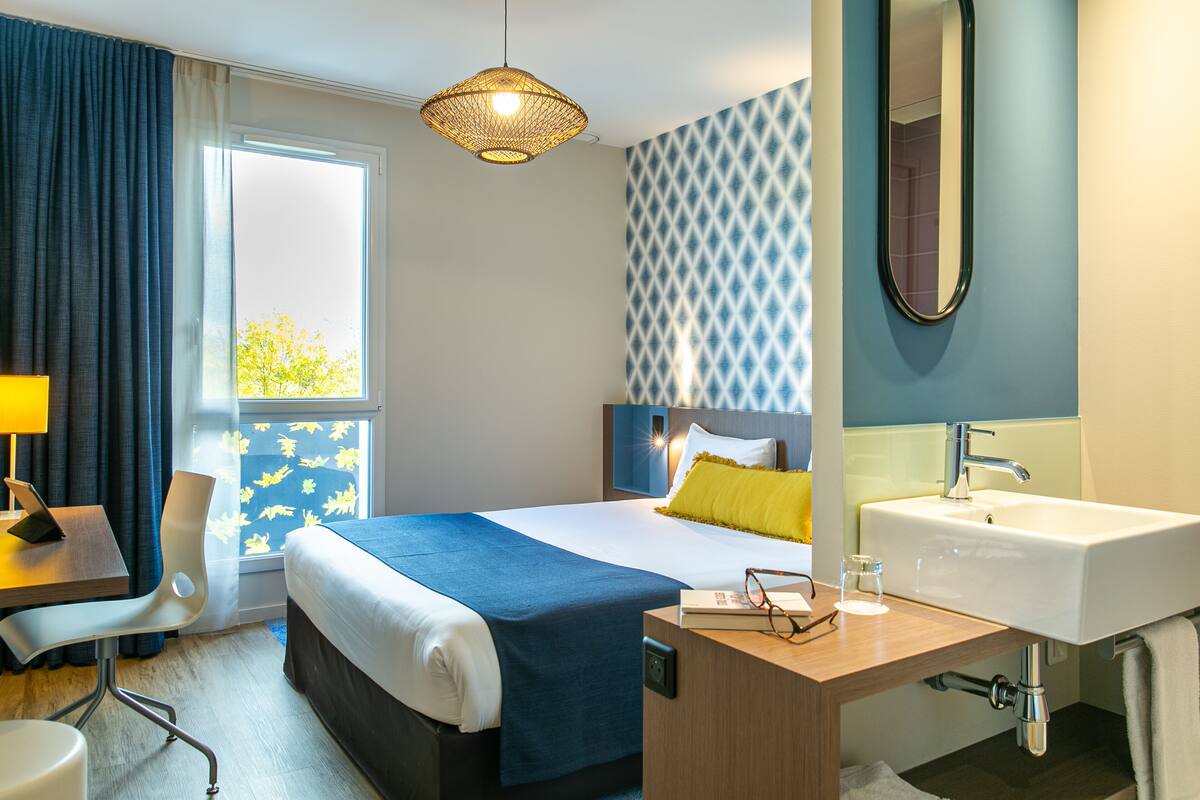 A hotel room with a bed, bedside table, lamp, chair, and a bathroom with a sink and mirror.