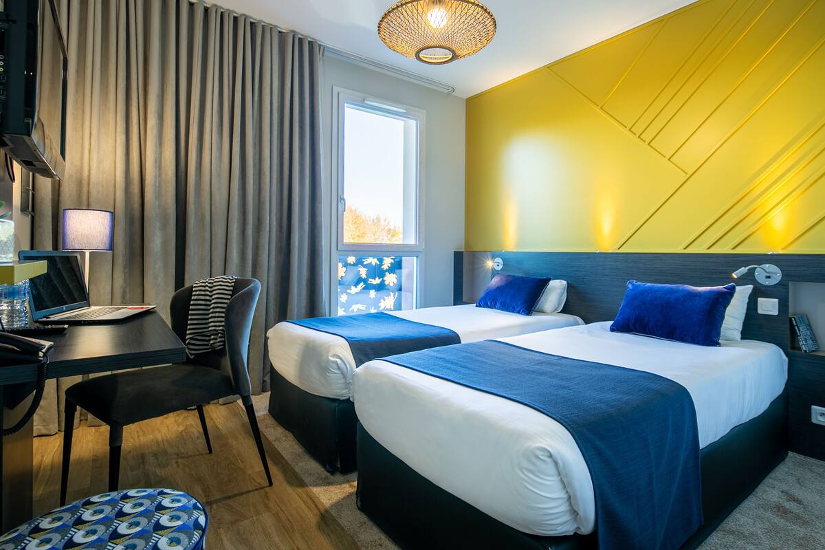 A hotel room with two beds, a desk with a laptop, a chair, and a small table with a lamp.