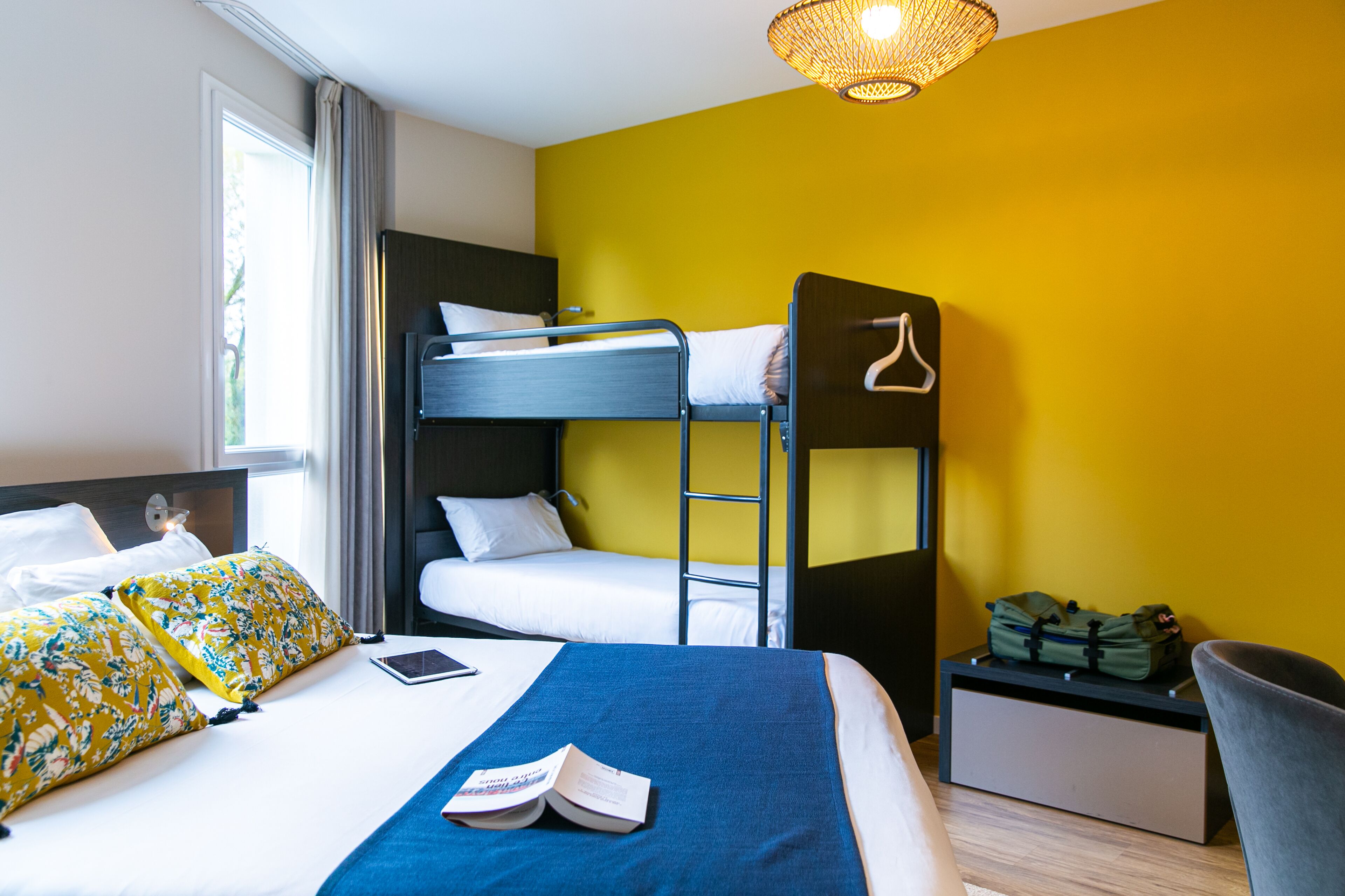A hotel room with a bunk bed, a desk with a chair, a small table with a bag, and a window with curtains.
