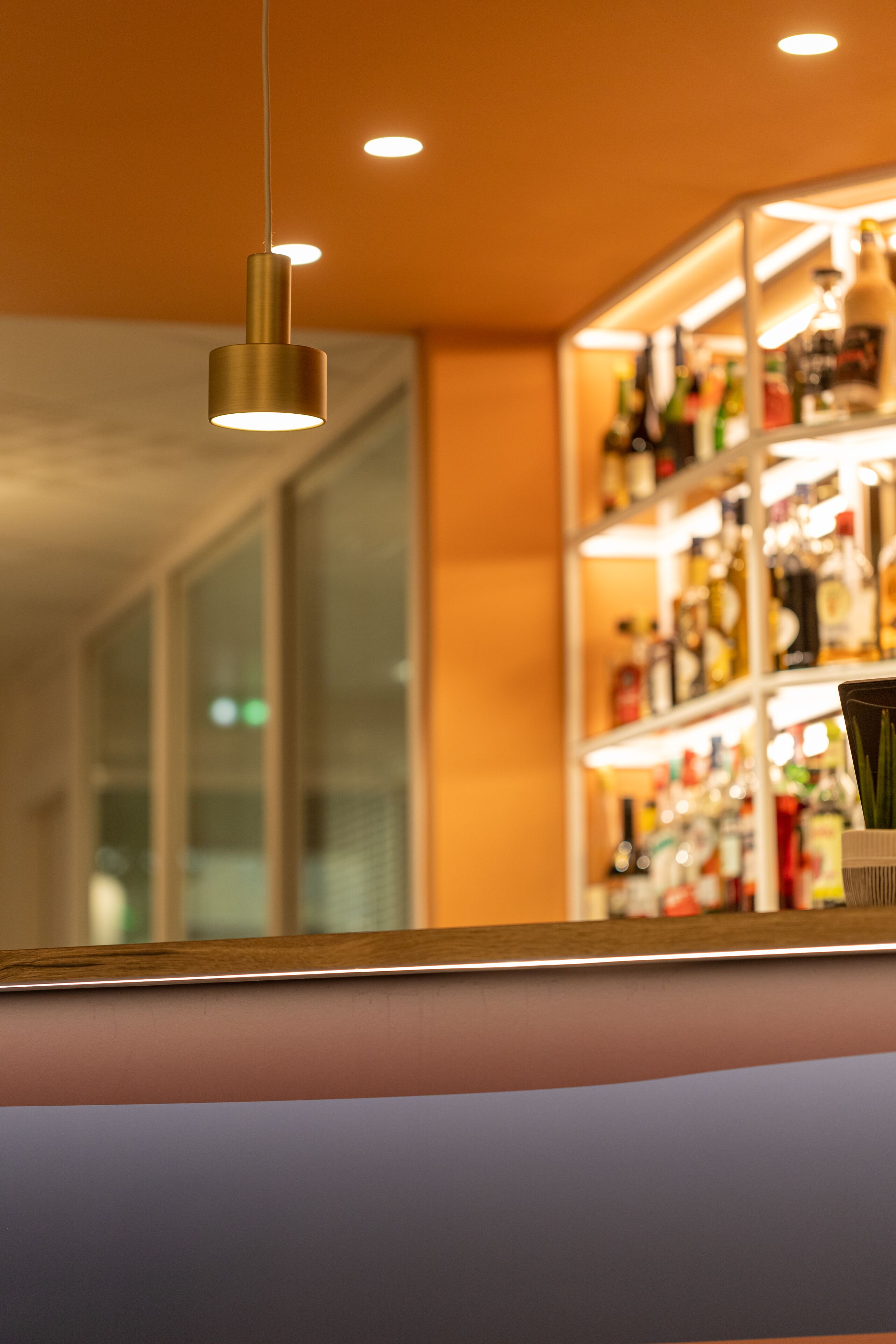 Una zona de bar con estantería llena de botellas, una lámpara colgante y un mostrador con superficie reflectante.