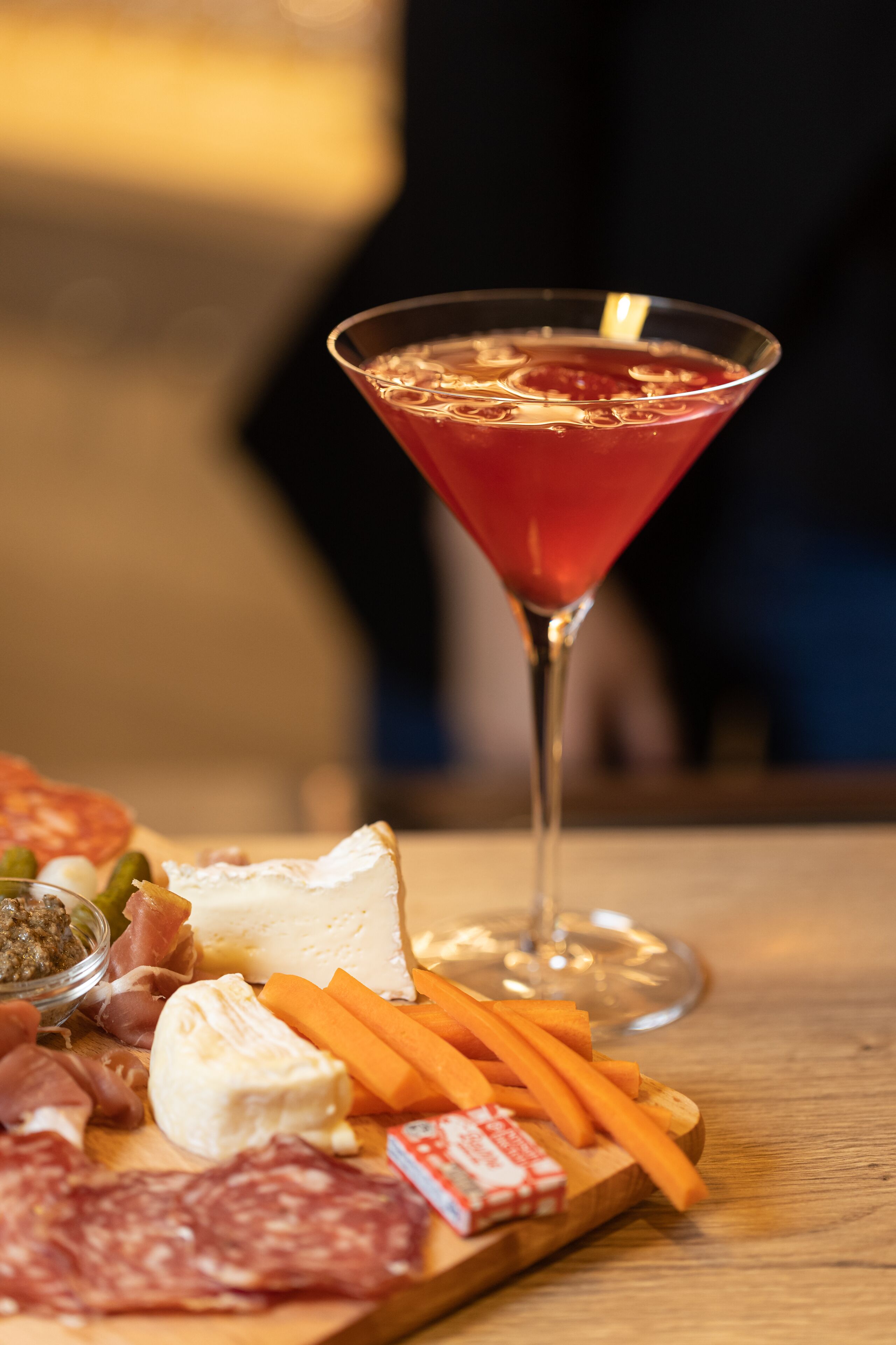 Una copa de martini con una bebida roja, una tabla de madera con diversos quesos, carnes y verduras.