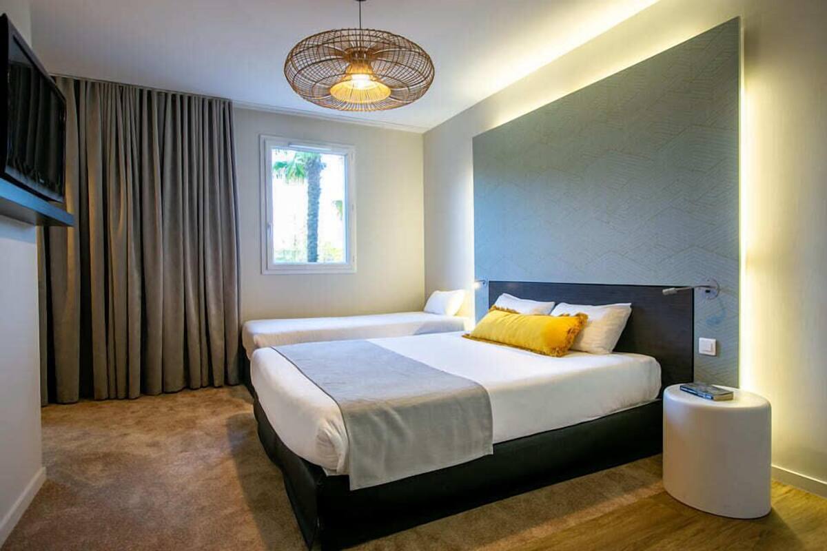 A modern hotel room with a large bed, a bedside table, a small round table, a ceiling fan, and a window with curtains.