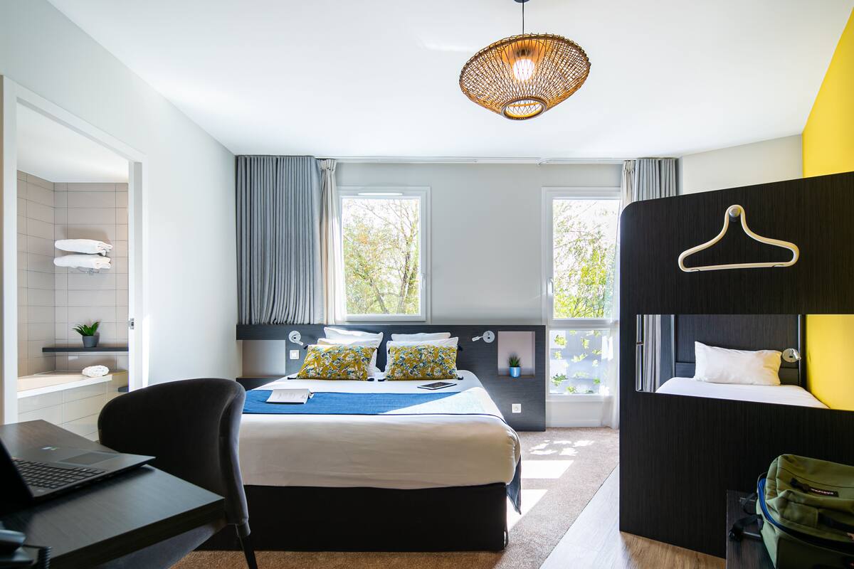 A modern hotel room with a bed, a desk with a laptop, a hanging light fixture, and a view of trees through the windows.