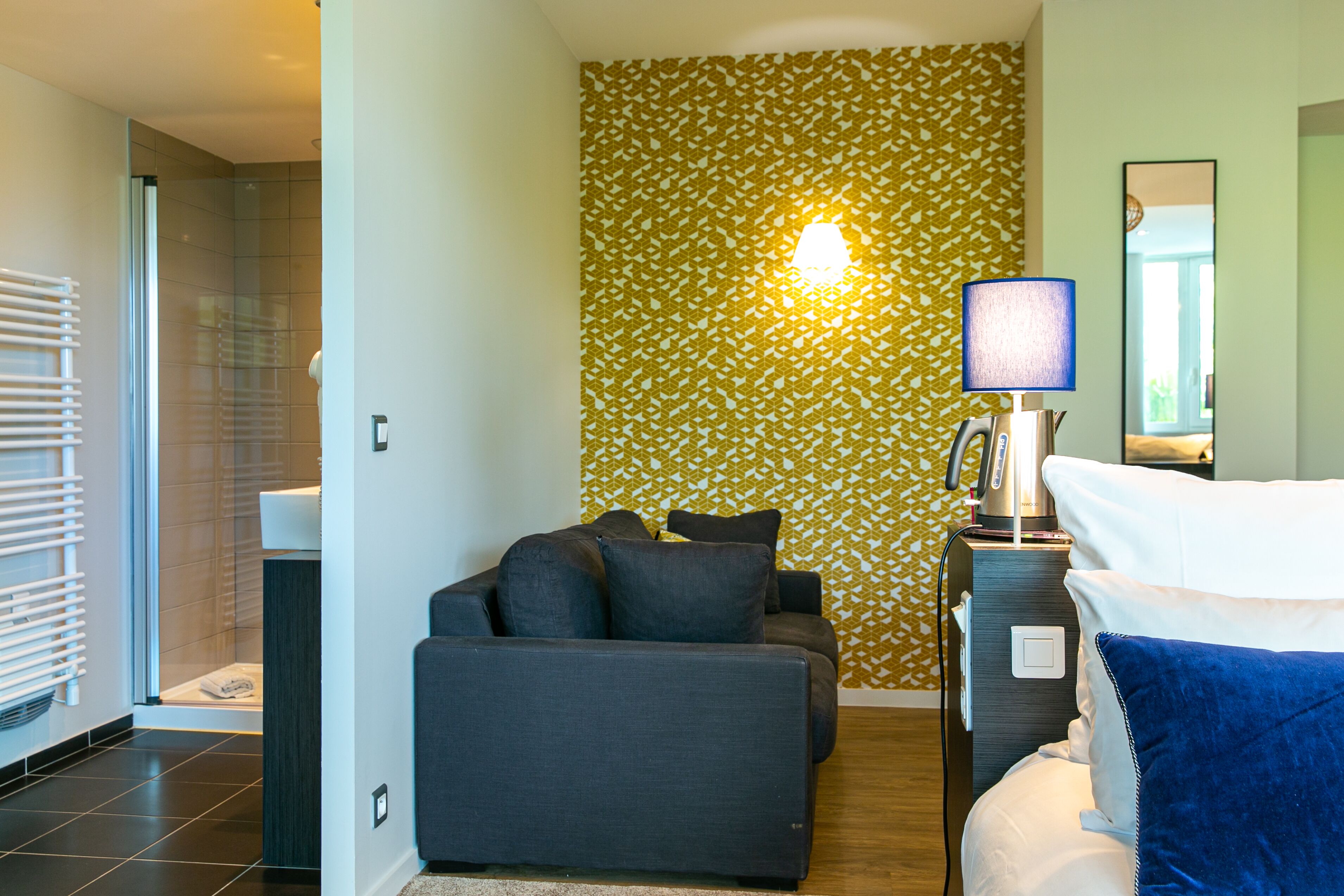 Una habitación de hotel moderna con sofá, una mesita de noche con lámpara, un espejo y un papel tapiz estampado.