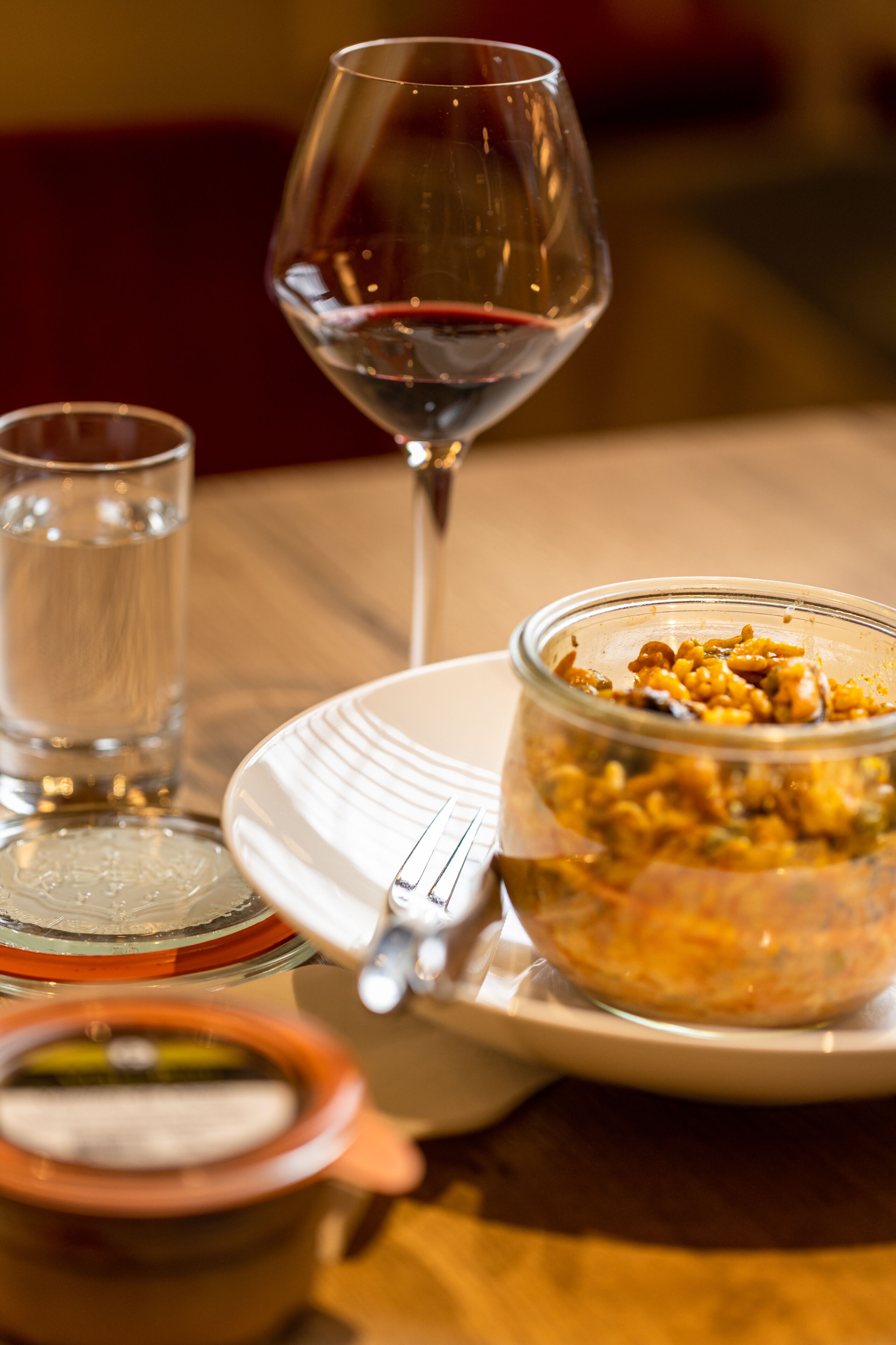 Una mesa de comedor con una copa de vino tinto, un vaso de agua, un plato con un tenedor y un tarro de comida.
