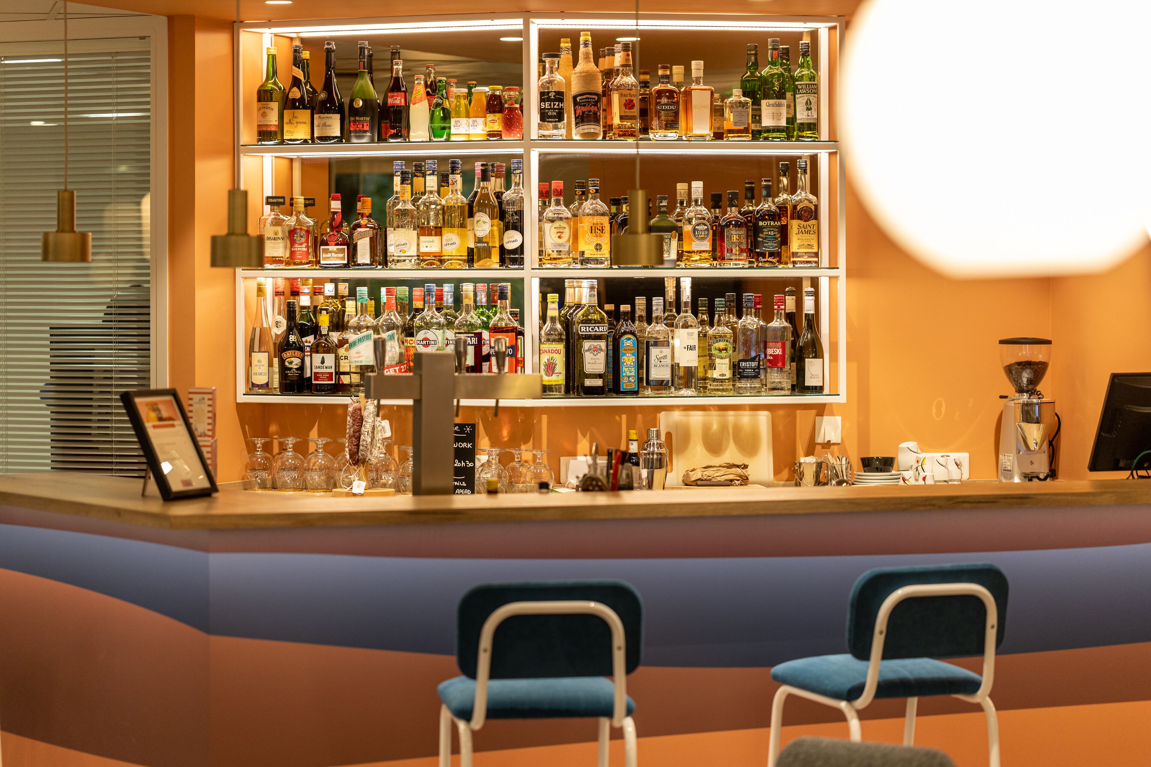 Zona de bar con estantes llenos de botellas de licor, dos sillas azules y una máquina de café.