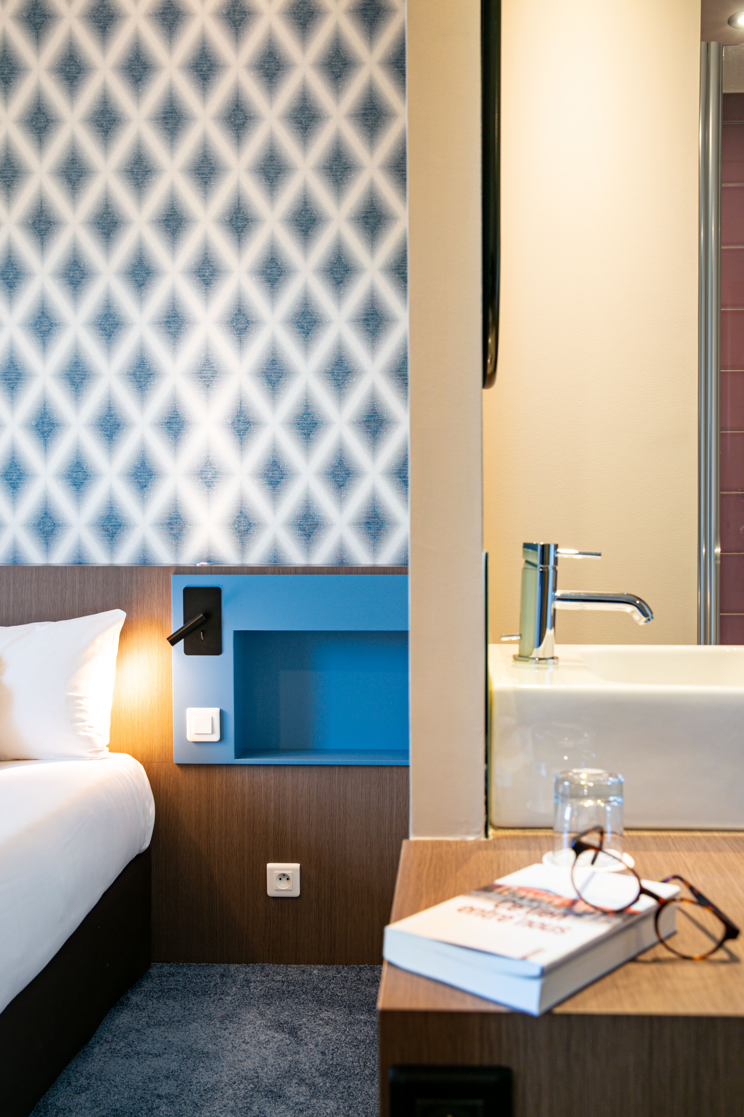 Ein Hotelzimmer mit einem ordentlich bezogenen Bett, einem Nachttisch mit Buch und Brillen, einer Wandleuchte sowie einem Badezimmer mit einem modernen Waschbecken und Spiegel.