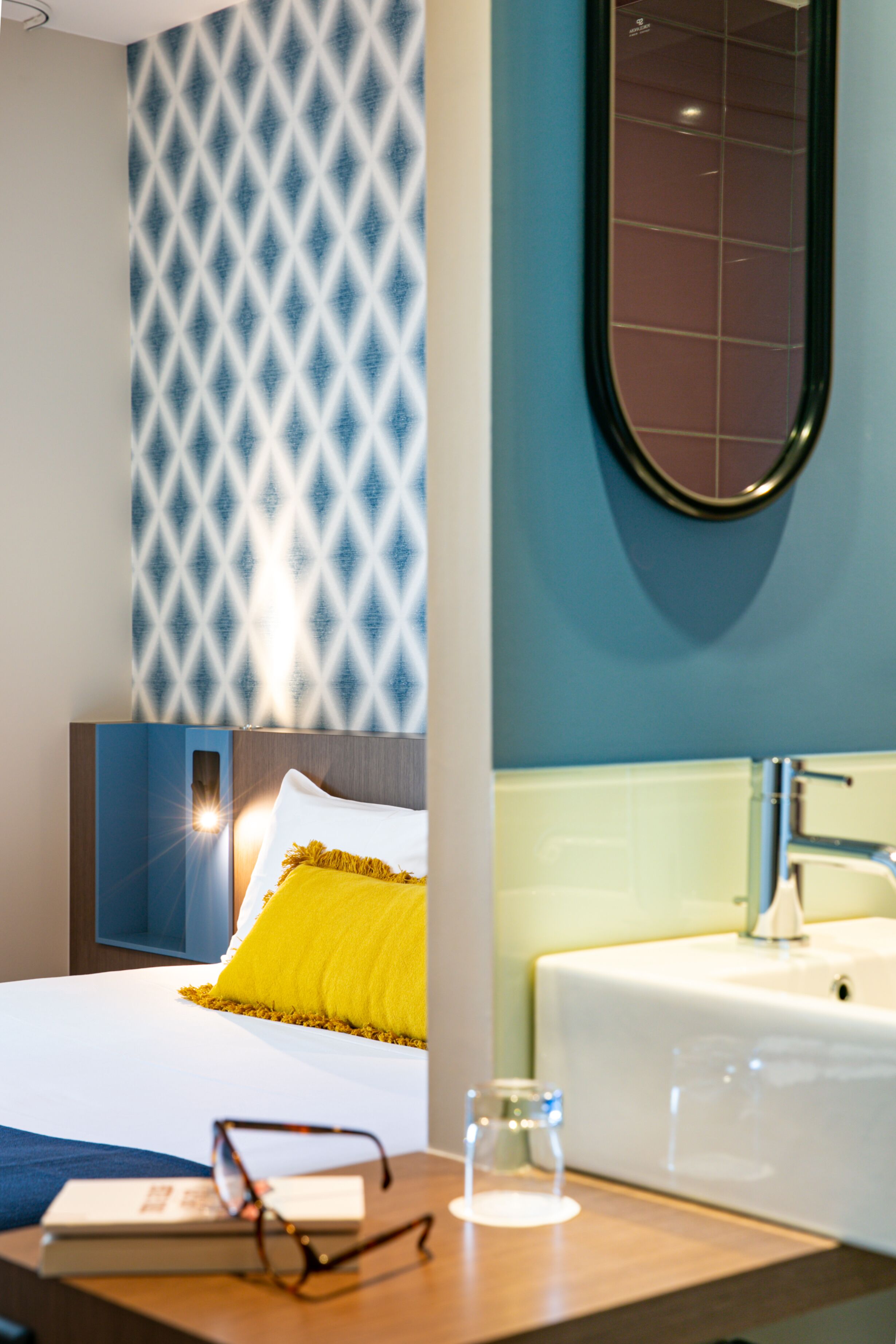 Habitación de hotel con cama, mesita de noche con vasos y un libro, baño con lavabo y espejo, y pared con diseño.