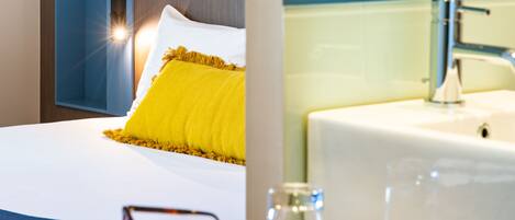 A hotel room with a bed, a bedside table with glasses and a book, a bathroom with a sink and mirror, and a patterned wall.